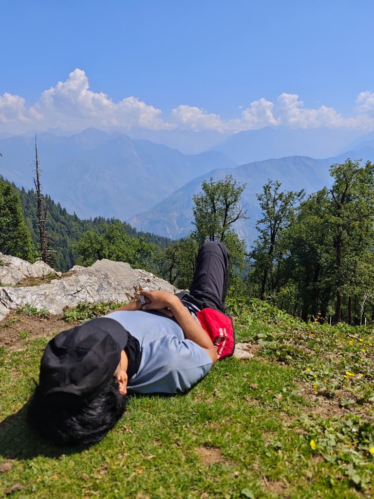 Vishwesh resting on the grass on a mountain ridge with the Himalayas in the background